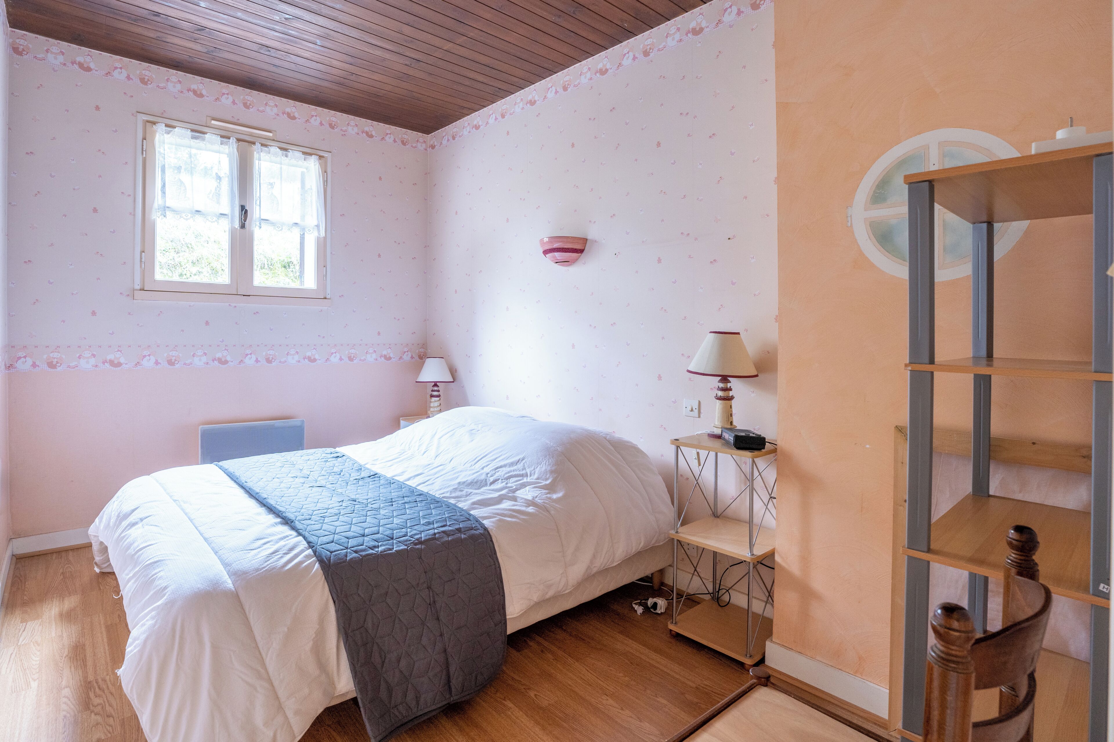 A bedroom with a bed, a nightstand, a lamp, a window, and a wooden shelf.