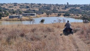Uma pessoa conduz uma motocicleta em um campo de grama seca, com um corpo d'água e edifícios ao fundo.