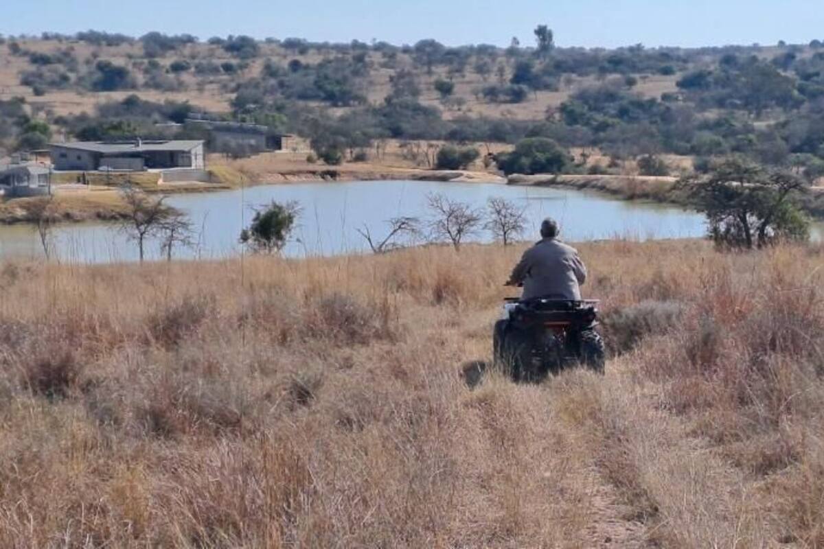 A person riding a motorcycle in a dry grass field with a body of water and buildings in the background.