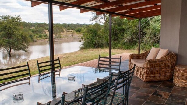 Uma mesa de jantar com tampo de vidro, cadeiras, um sofá de vime e vista para um lago.