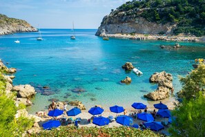 Ein Küstenstrand mit klarem türkisblauem Wasser, Felsformationen und einem Sandbereich mit blauen Sonnenschirmen.