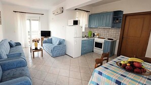 Een woonkamer met een bank, een kleintje tafeltje met een vaas, een keuken met blauwe kastjes, een eettafel met fruit, en een deur.