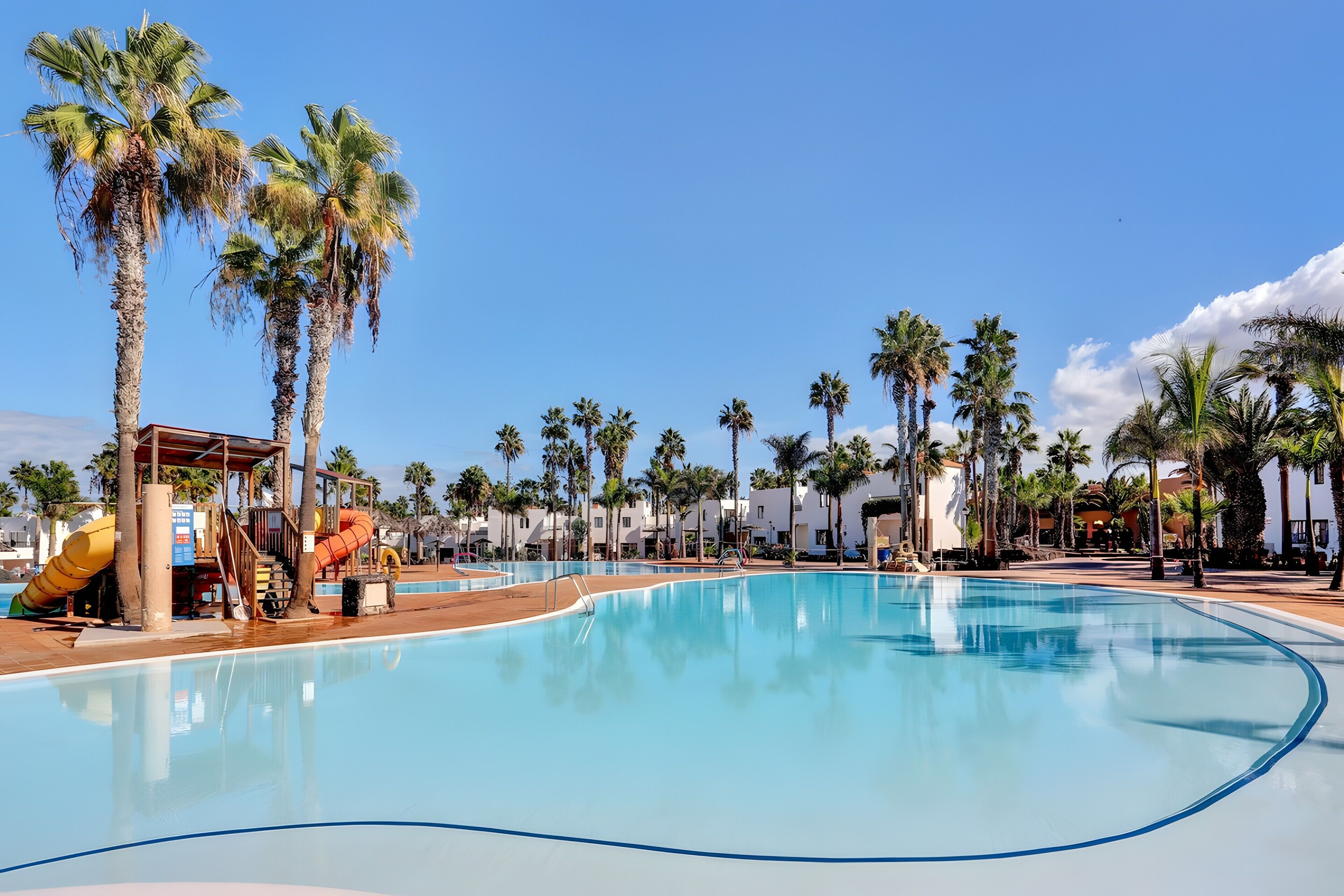 A swimming pool with palm trees, a water slide, and a building with a red roof.