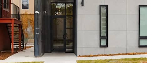 A modern building entrance with a glass door, a staircase, and a small outdoor area with grass.