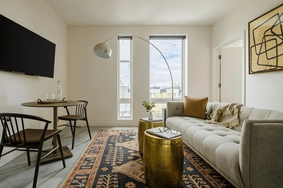 A modern living room with a sofa, a dining table with chairs, a TV mounted on the wall, and large windows.
