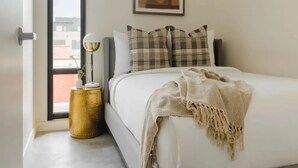 A bedroom with a bed, pillows, a throw blanket, a lamp, and a painting on the wall. - The Artesian (Denver)
