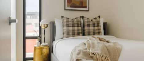 Un dormitorio con cama, almohadas, una manta decorativa, una lámpara y un cuadro en la pared.