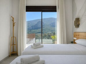 Een hotelkamer met een bed, een houten kapstok, een balkon met uitzicht op bergen en bomen, en een groot raam met gordijnen.