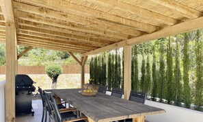 Een terras buiten met een houten tafel, stoelen, een barbecue en een stenen muur.
