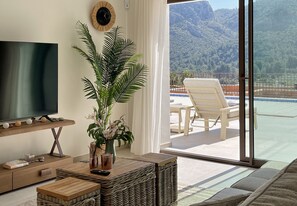 Ein Wohnzimmer mit Fernseher, einer Pflanze, einem Holztisch und Blick auf eine Berglandschaft durch eine Glastür.