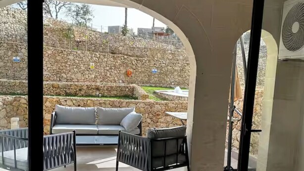 A modern interior with a sliding glass door leading to an outdoor seating area with a stone wall backdrop. - Luxury 3BR Escape - Pools and Gardens (Qala)