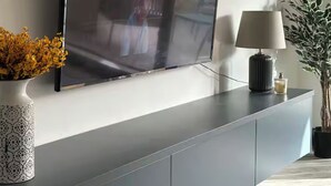 A modern living room with a flat-screen TV mounted on a grey cabinet, a lamp, and a potted plant. - Luxury 3BR Escape - Pools and Gardens (Qala)