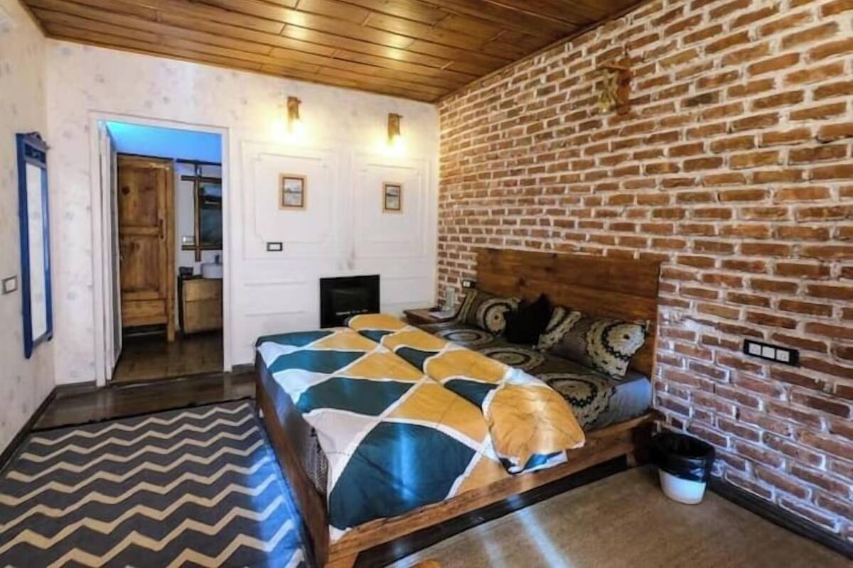 Una habitación con techo de madera, pared de ladrillo, cama con ropa de cama estampada, un televisor y una puerta que da a otra habitación.