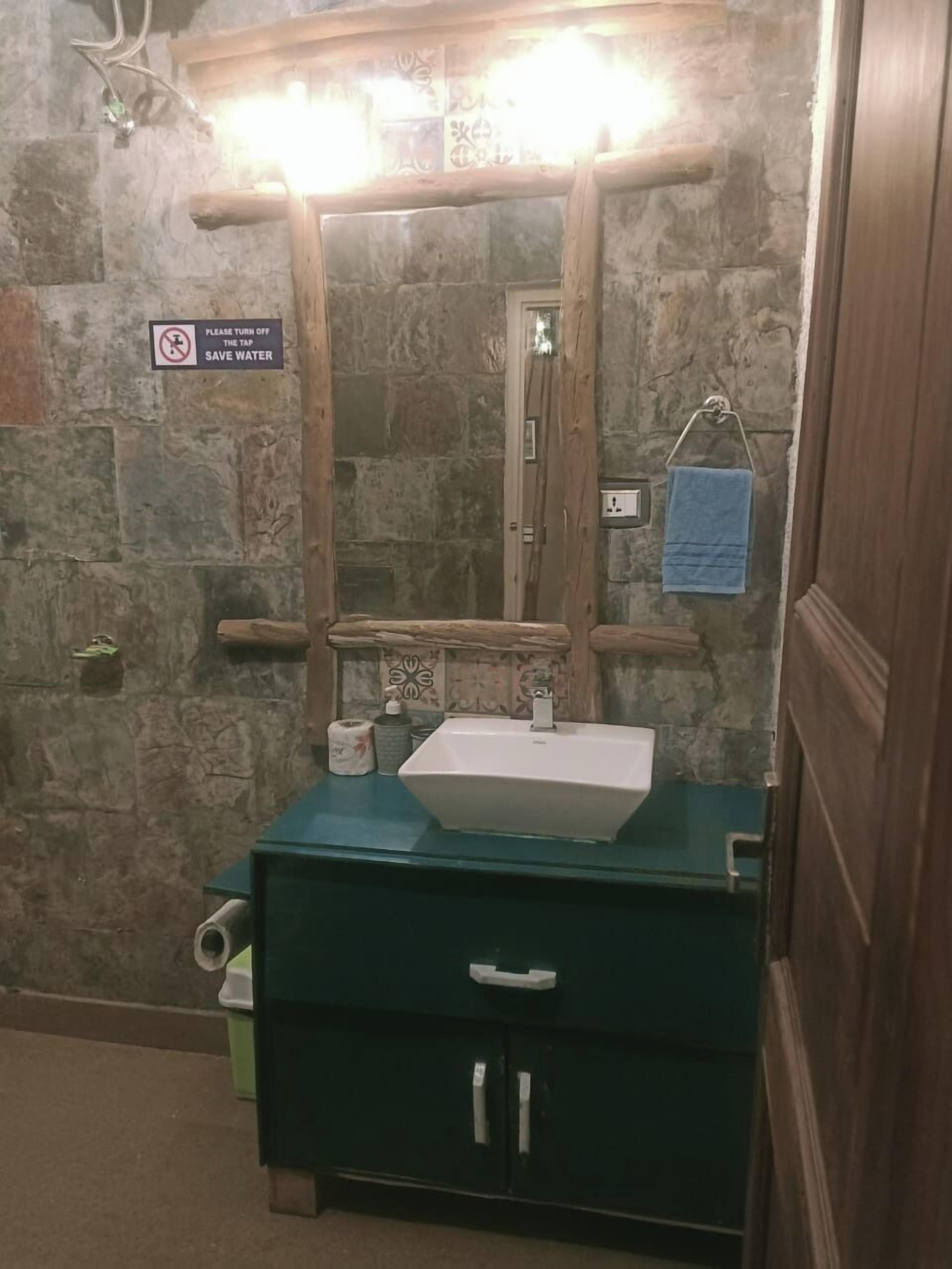 A bathroom with a stone wall, a wooden vanity, a mirror with lights, a towel rack, and a 'Please turn off the tap save water' sign.