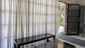 Ein modernes Wohnzimmer mit Glastisch und Sofa, ein großes Fenster mit transparenten Vorhängen und eine Glastür, die nach draußen führt.