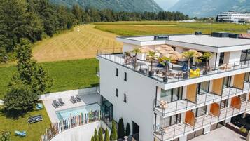 Un edificio moderno con terraza en la azotea, piscina y vistas a un campo verde y montañas.