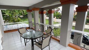 A covered balcony with a glass table and chairs, white tiled floor, and a view of greenery and buildings. - 3-bedroom luxurious house in Kingston with pool, private parking free WiFi (Kingston)
