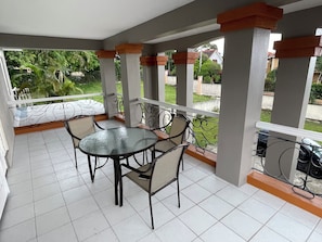 Un balcón techado con una mesa y sillas de vidrio, piso de baldosas blancas y vista a zonas verdes y edificios.