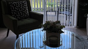 Ein Zimmer mit einem Glastisch, einer gepolsterten Couch mit einem gemusterten Kissen und Blick durch einen weißen Zaun auf einen Außenbereich mit Stühlen.