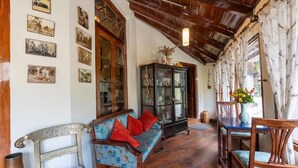 A room with a wooden ceiling, a blue patterned sofa with red cushions, a wooden cabinet, and a dining table with chairs. - Alaya Stays The Hive Cottage (Nainital)