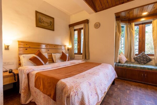Une chambre à coucher avec une tête de lit en bois, un lit recouvert d’une couverture marron, une table de chevet, une fenêtre avec des rideaux et un banc agrémenté de coussins.