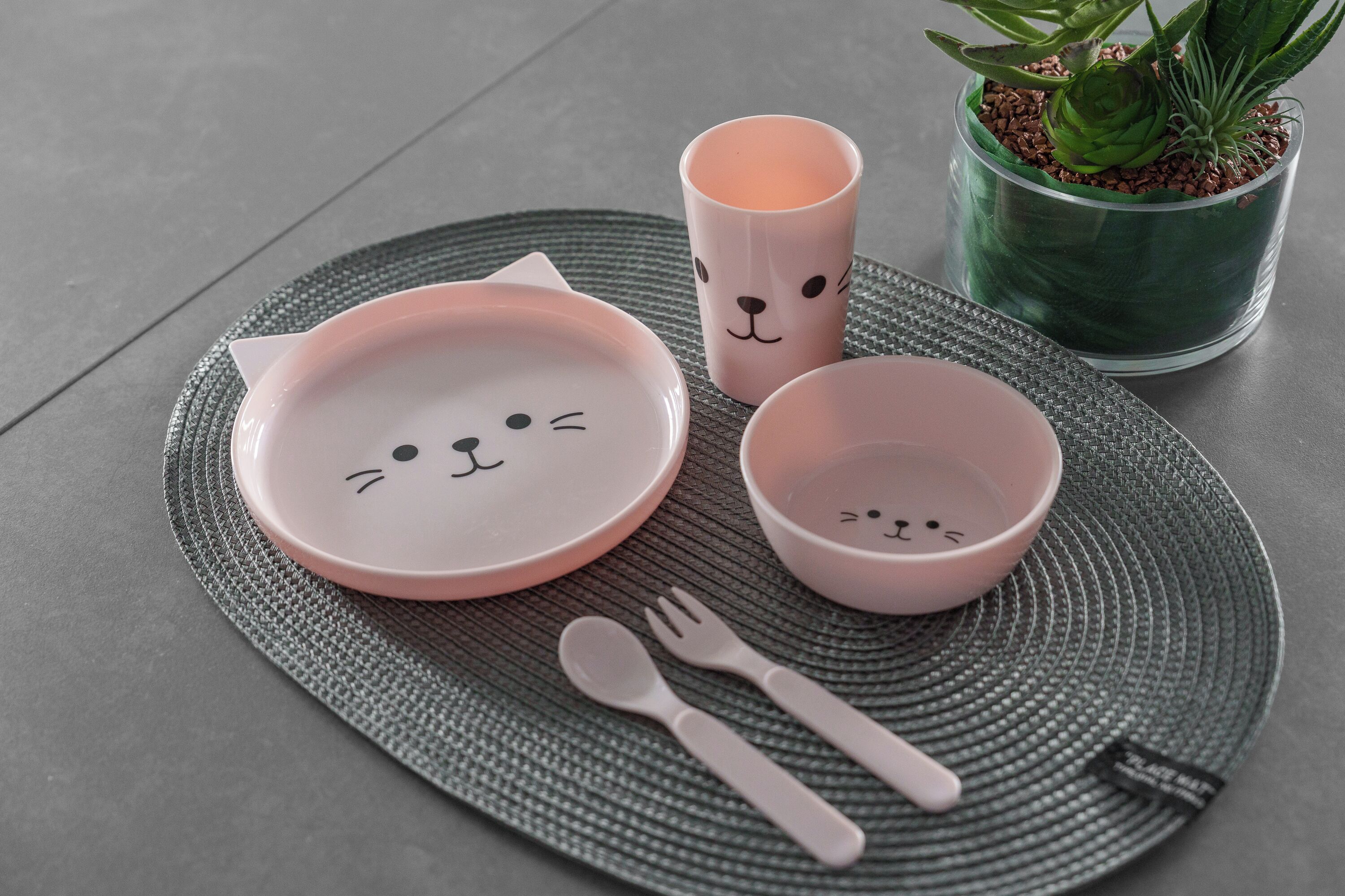 Et bord dækket med en tallerken med katte motiv, en kop, en skål og bestik.