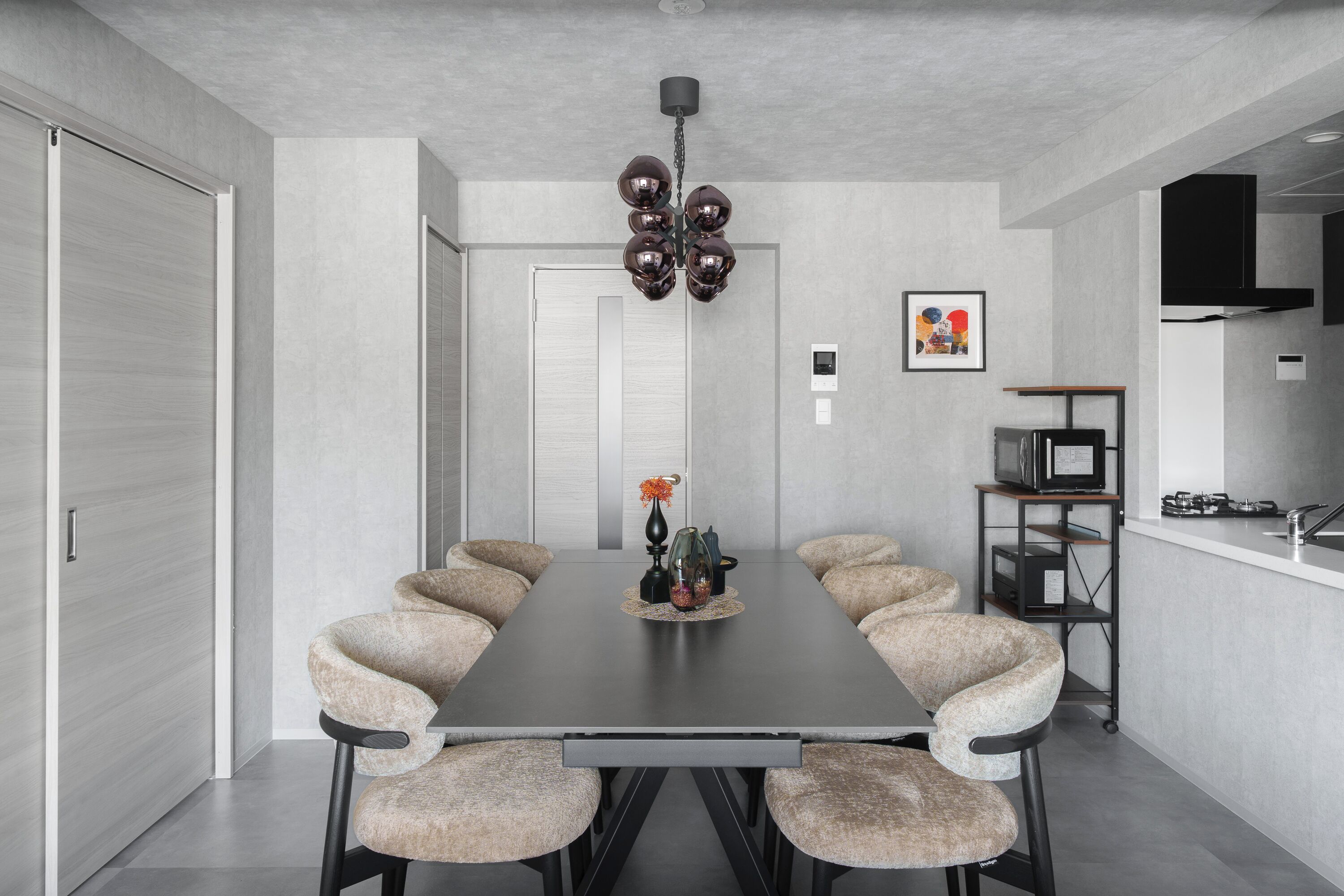 A modern dining area with a long table, six chairs, a chandelier, and a kitchen in the background.