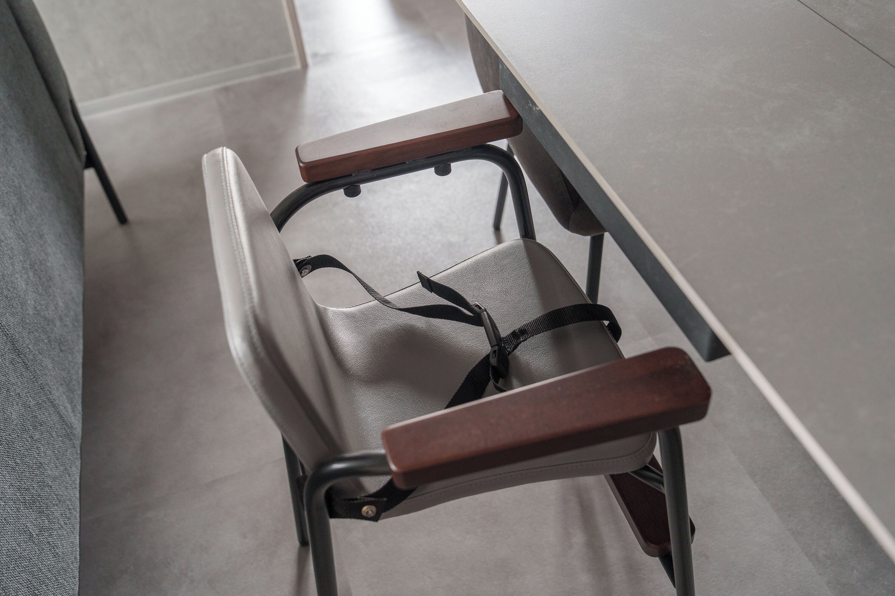 Una silla con respaldo y apoyabrazos de madera, unida a una estructura metálica, en una habitación con piso de baldosas.