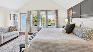 Ein Schlafzimmer mit einem großen Bett, einem Nachttisch, einer Lampe, einem Fenster mit Vorhängen, einem Deckenventilator und Blick ins Freie.