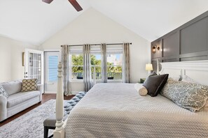 A bedroom with a large bed, a nightstand, a lamp, a window with curtains, a ceiling fan, and a view of the outdoors. - Recently Renovated | Ocean View 3 BR Home | Steps To The Beach | Resort Access (Isle of Palms)