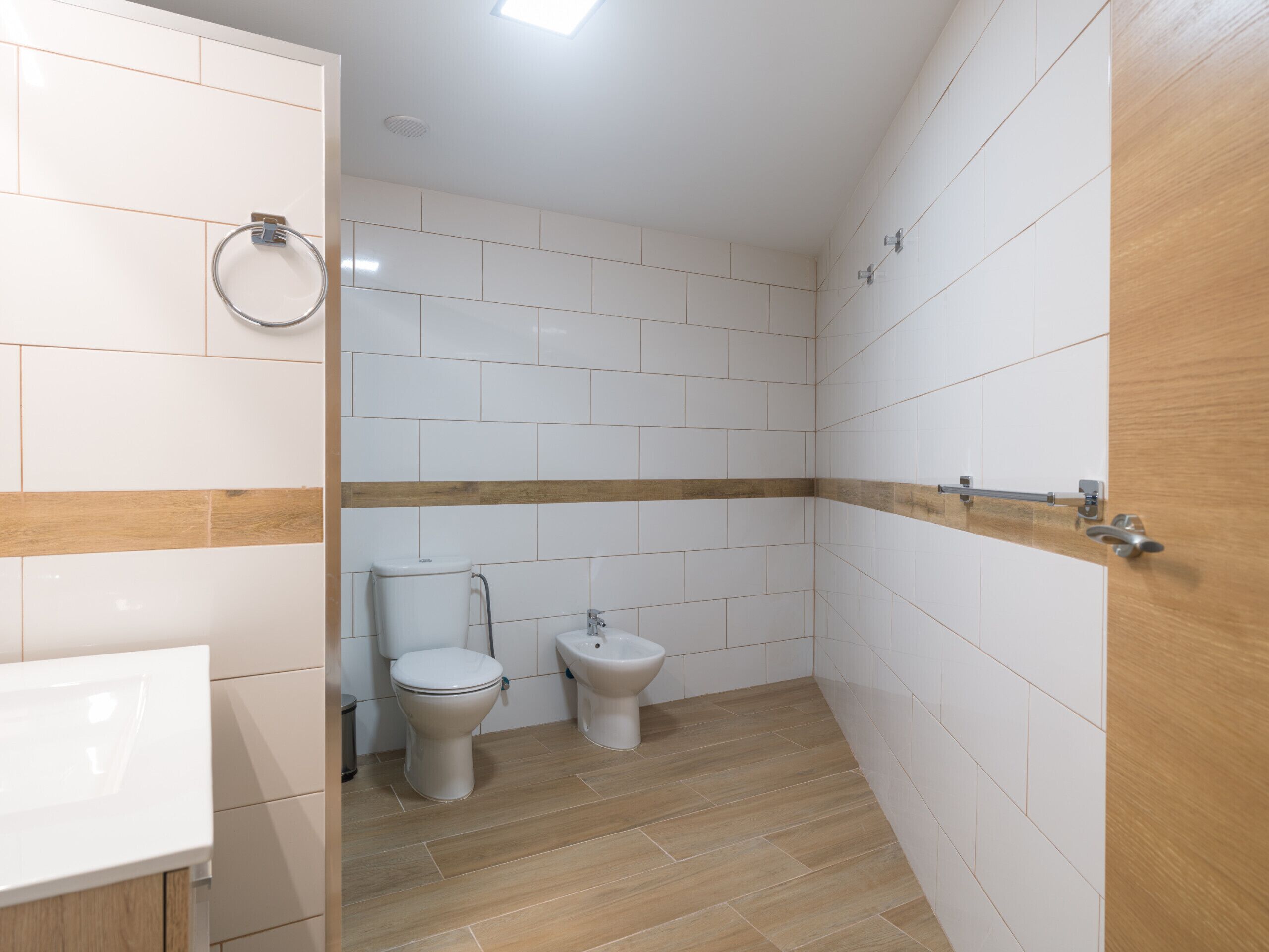 A bathroom with white tiles, a wooden floor, a toilet, a bidet, and a sink.