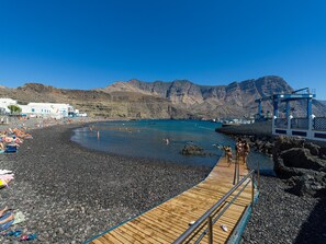 A beach with a wooden pier leading to a rocky shore, a clear blue sky, and mountains in the background. - Aguadulce Agaete Bajo (Agaete)