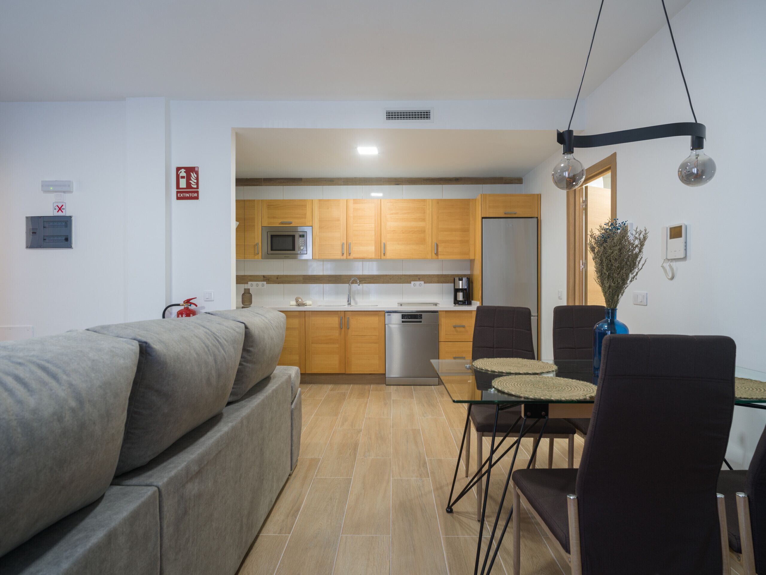 Een moderne keuken met houten kastjes, een magnetron en een koelkast. Er is een eettafel met stoelen en een vaas met gedroogde bloemen. Een bank bevindt zich op de voorgrond.