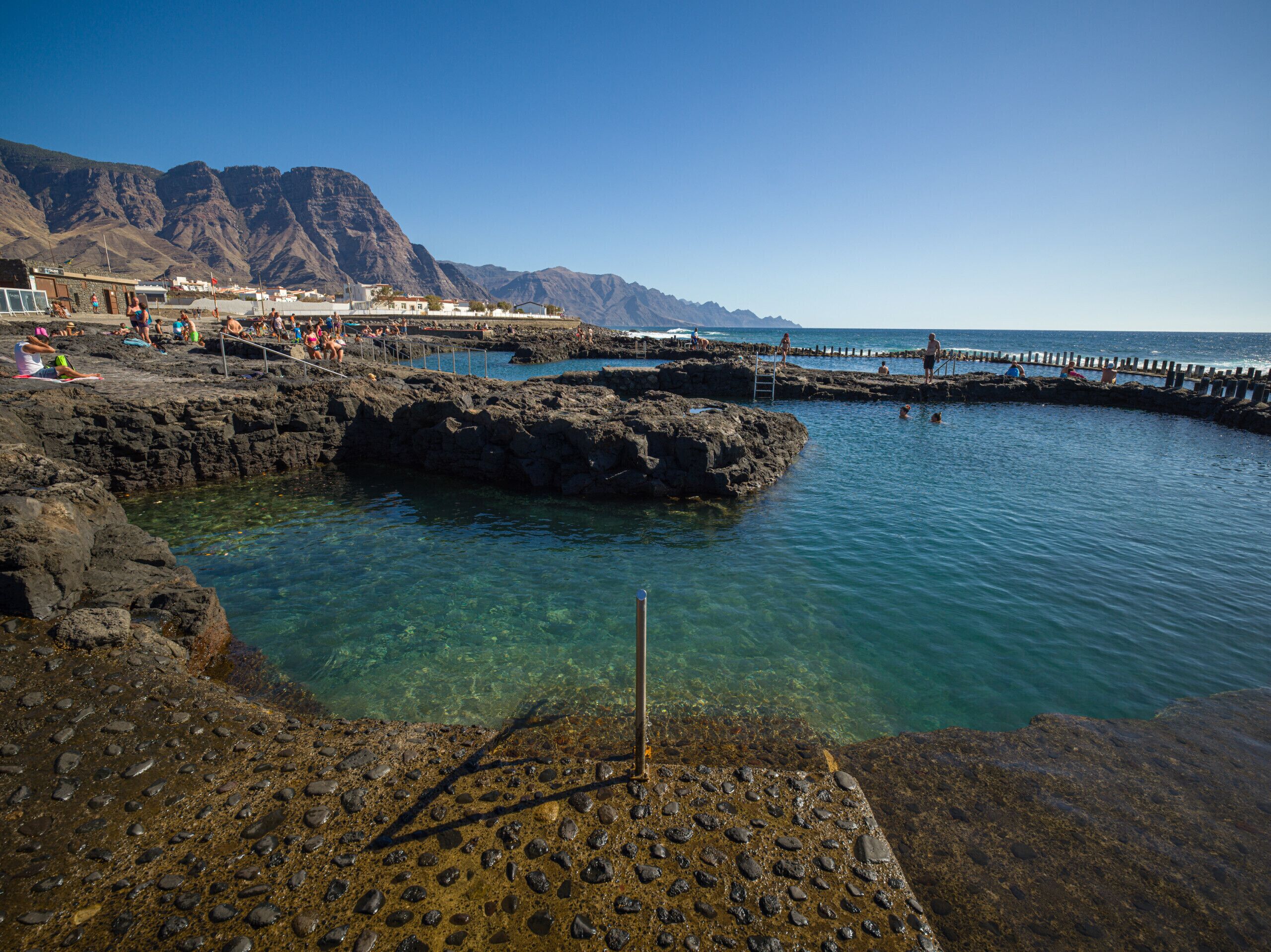 A rocky coastal area with clear water, a wooden pier, and mountains in the background.