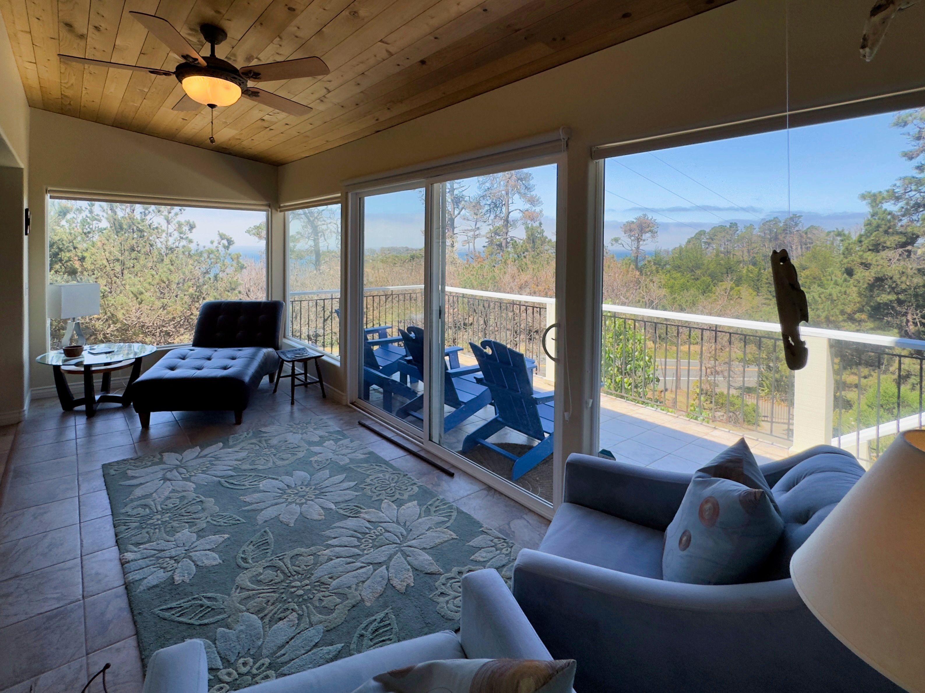 Une pièce dotée d’un ventilateur de plafond, d’un canapé, d’une table avec des chaises, d’un tapis et de grandes fenêtres offrant une vue sur les arbres et les montagnes.
