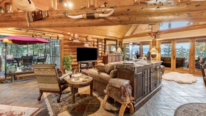Interior de una cabaña de troncos con chimenea, muebles de madera, televisor y comedor con vistas al exterior.