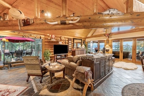 A log cabin interior with a fireplace, wooden furniture, a television, and a dining area with outdoor views.