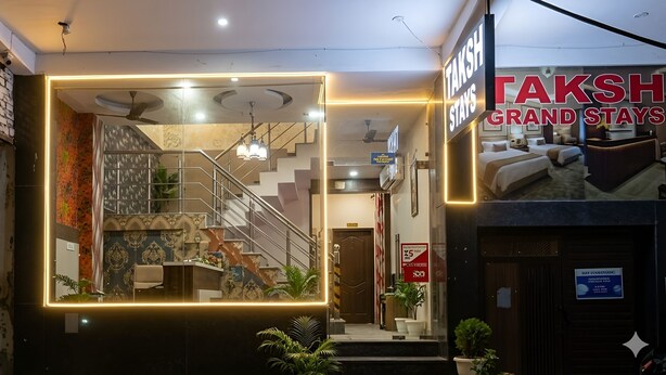 Interior de hotel con una escalera, una recepción y un cartel que dice 'TAKSH GRAND STAYS'.