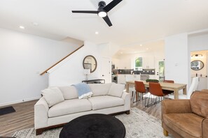 Una sala moderna con un sofá, una mesa de centro, un comedor con sillas, una cocina al fondo y un ventilador de techo.