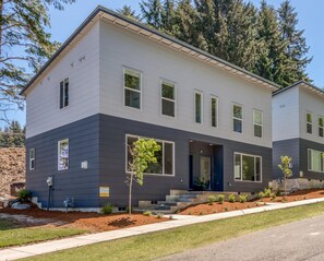 Une maison moderne à deux étages, dotée d’une façade sombre, de grandes fenêtres et d’un jardin bien entretenu avec de jeunes arbres.