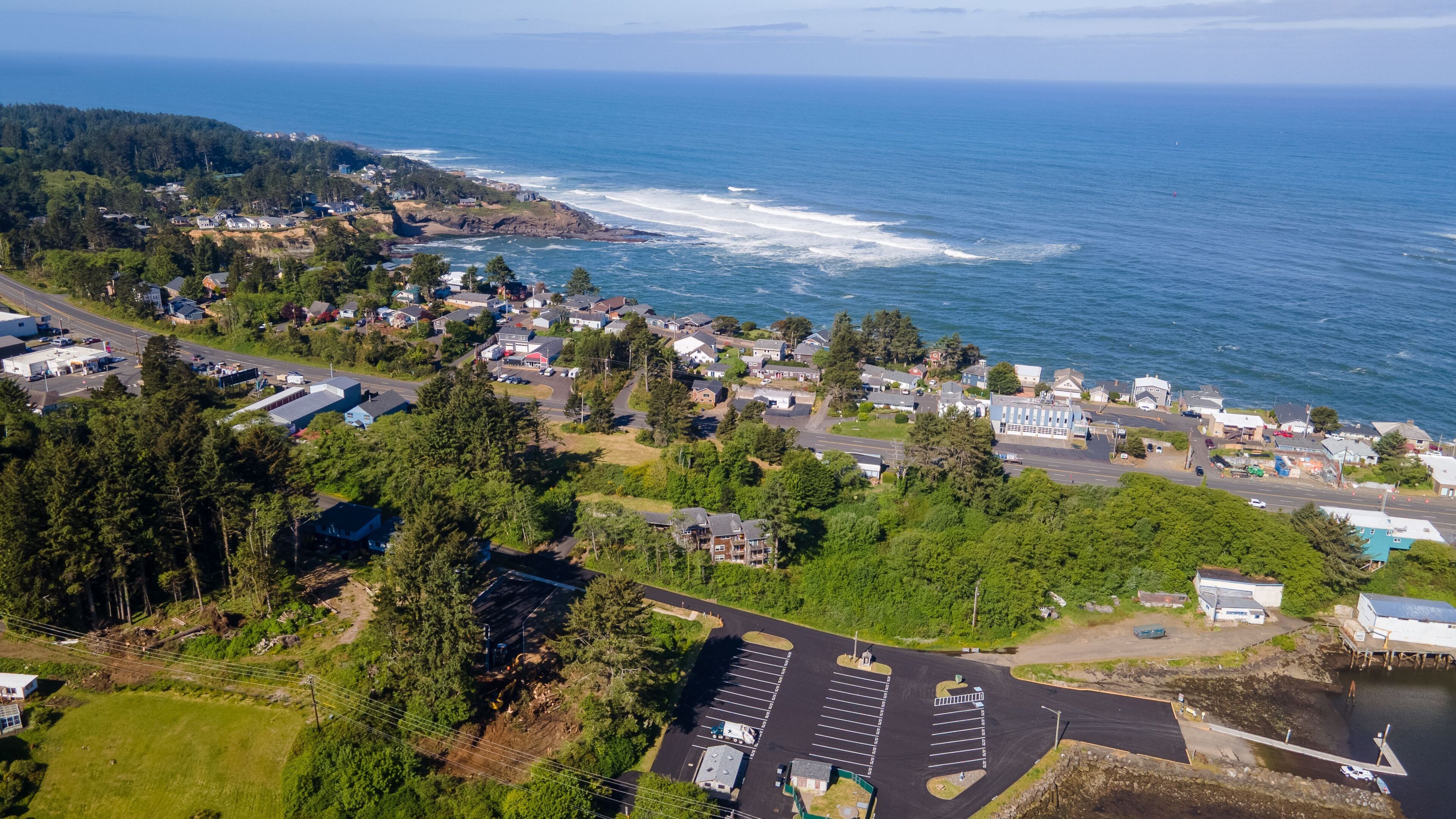 An aerial view of a coastal town with residential buildings, a parking lot, and a road leading to the beach.