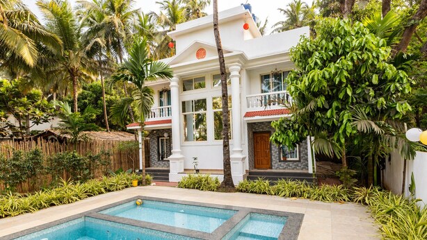 A pool with a tiled deck, a white building with a red door, and lush greenery.