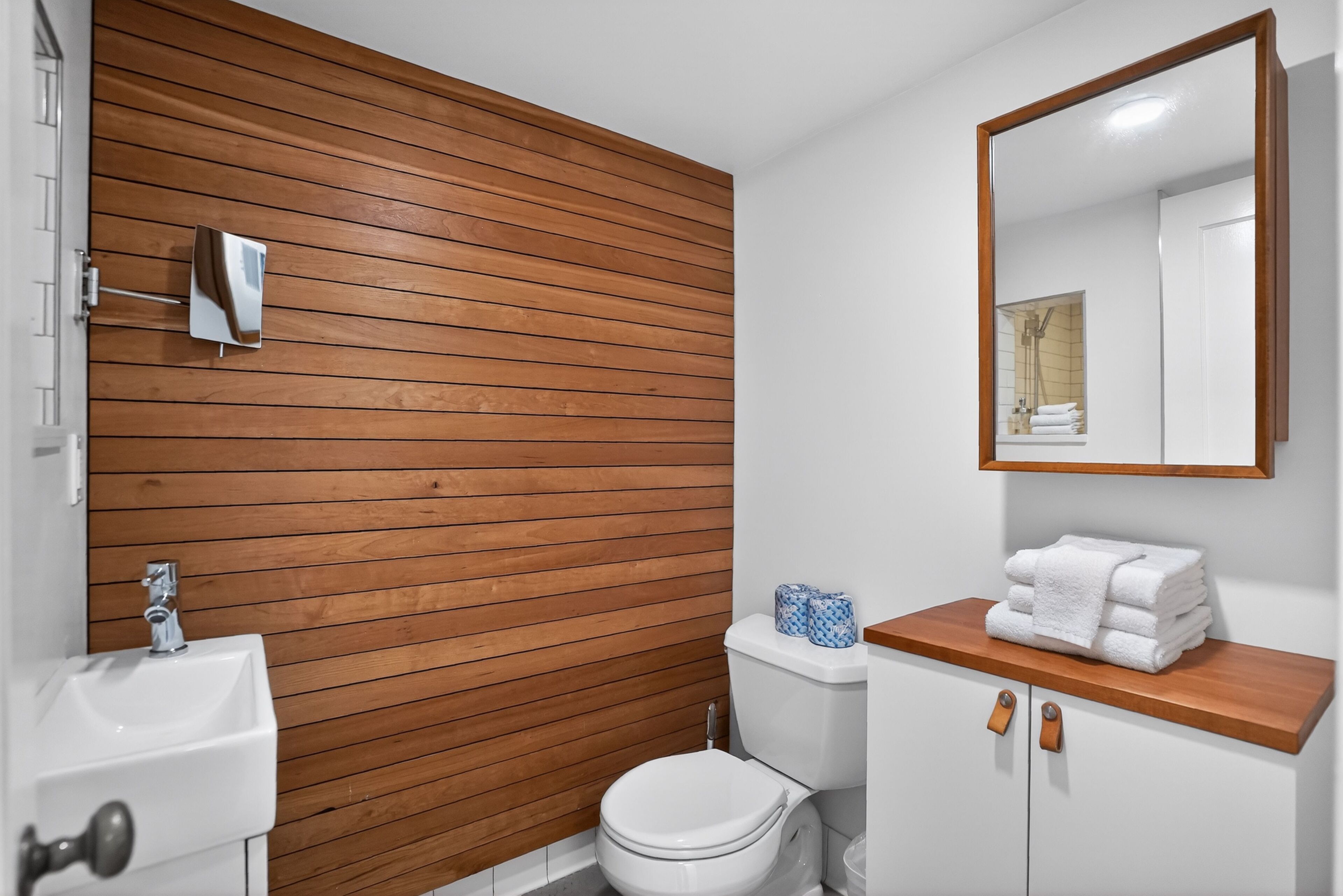 A modern bathroom with a wooden panel wall, a white sink, a toilet, and a wooden vanity with a mirror.