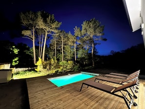 Une terrasse en bois avec une piscine, des chaises longues et de grands arbres en arriĂšre-plan.