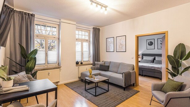 Ett modernt vardagsrum med en soffa, ett matbord och ett soffbord. Det finns två fönster med gardiner, ett element och en dörr som leder till ett sovrum.