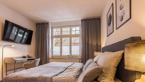Ett sovrum med en säng, ett skrivbord, en stol, en tv, ett fönster med gardiner och konst på väggen.