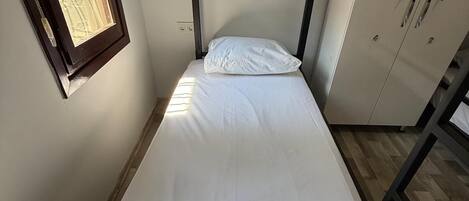 Una cama individual con colchón y almohada blancos, una ventana pequeña y un armario de dos puertas.