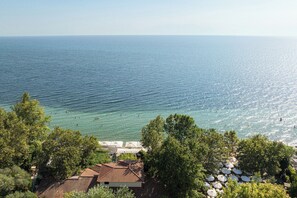 Une vue sur la côte, avec un ciel dégagé, une mer calme et un bâtiment doté d’un toit en tuiles, ainsi que des arbres au premier plan.