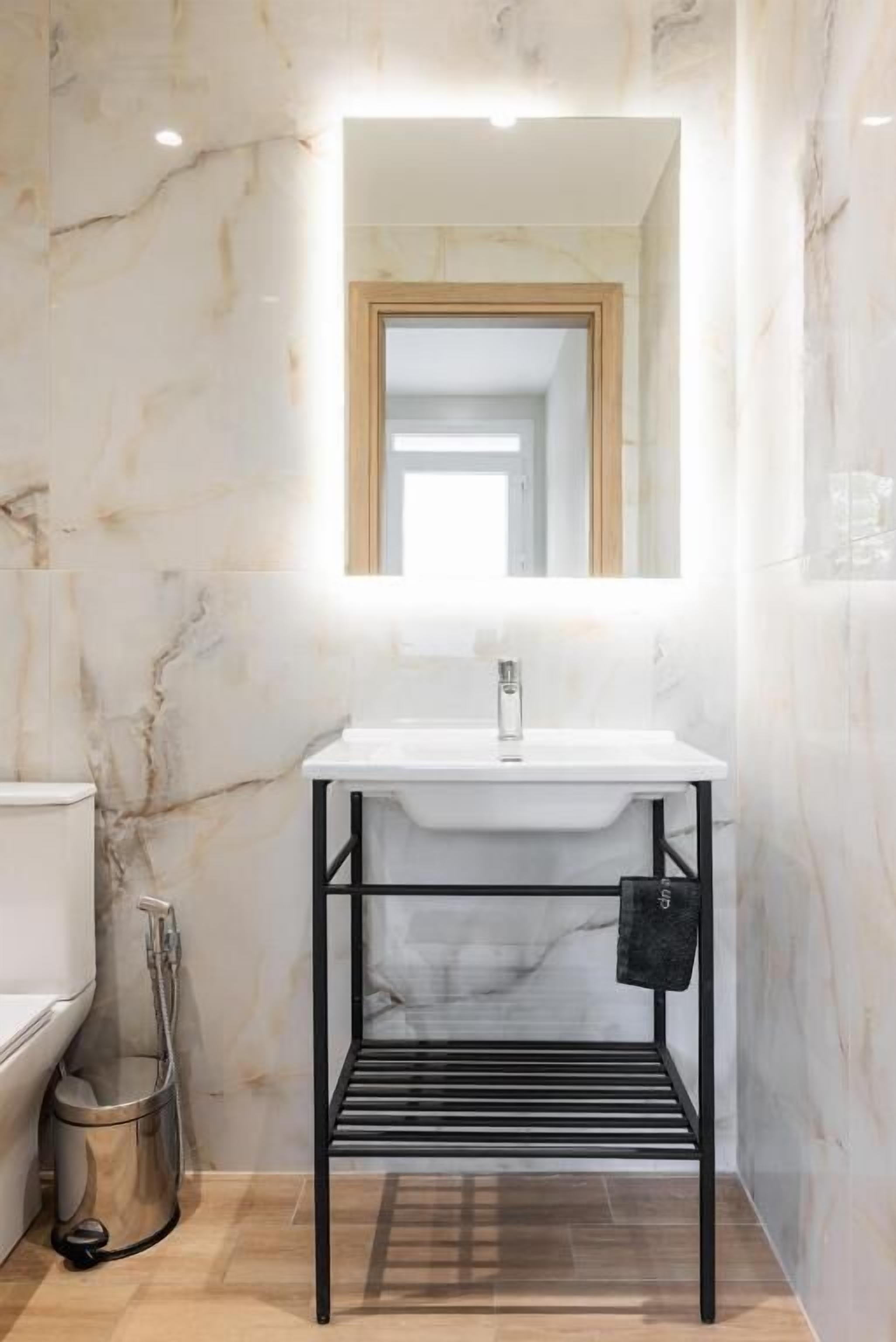 Un baño con pared de mármol, un lavamanos blanco, un soporte de metal negro y un espejo.