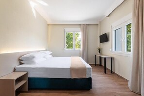Un dormitorio con cama, mesita de noche, una pequeña mesa con una planta, televisor montado en la pared y ventana con cortinas.